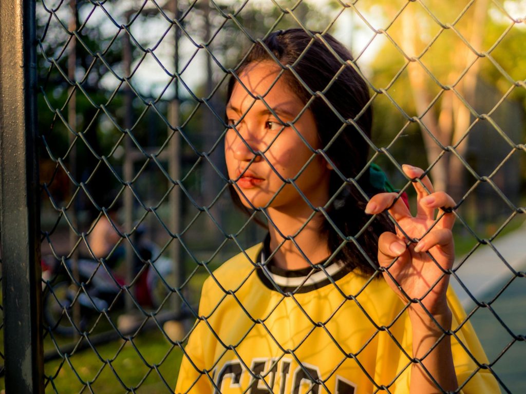 A young woman in a yellow shirt stands thoughtfully behind a chain-link fence, with sunlight casting shadows on her face. She holds onto the fence with one hand, while greenery and a blurred figure are visible in the background.